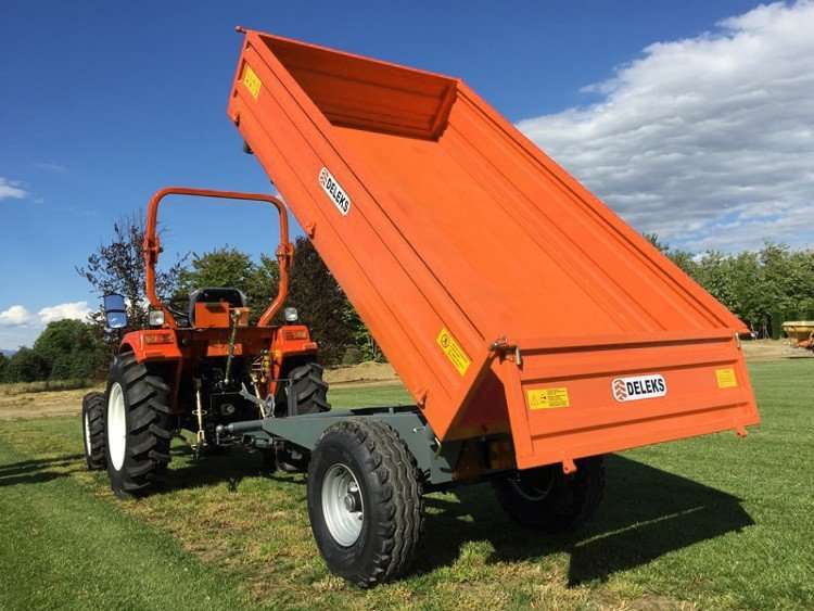 Oranž traktor on kinnitatud Kallutav käru RM-14-T3 kalluriga haagise külge, mille lava on tõstetud õues muru, puude ja sinise taeva taustal. Haagisel on Delens- ja CE-märgised.