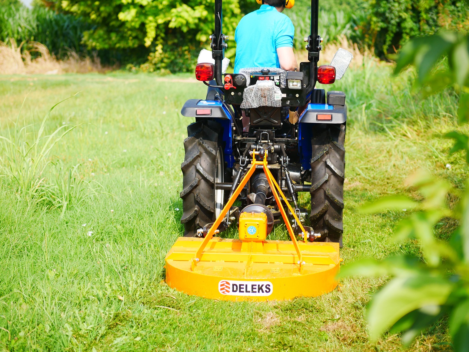 Inimene sõidab sinise traktoriga Deleks Tasandusniiduk BUGGY-100 niidukiga, niites rohtu kitsal, kõrghaljastusega ümbritsetud rajal. Foto on tehtud traktori tagant.