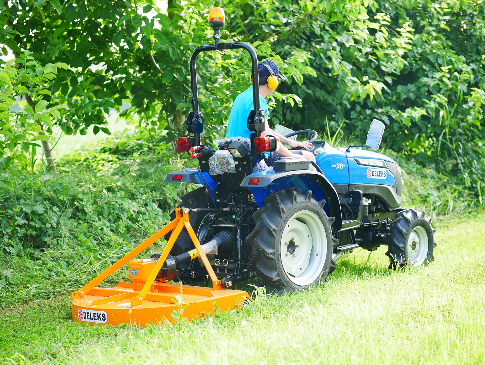 Inimene juhib sinist kompakttraktorit koos Deleks Tasandusniiduk BUGGY-100 niidukiga, niites rohtu mööda rohelist, puiduga piiratud ala, kandes kõrvaklappe ja mütsi.