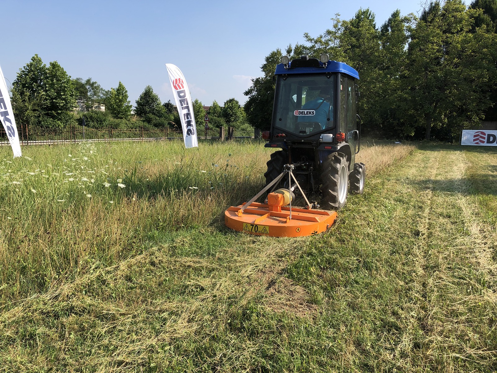Traktor Deleks tasandusniidukiga BUGGY-120 oranžikas niiduk lõikab põllul kõrget rohtu, vasakul kaks valget lippu, millel on logod ja tekst. Taustal on puud ja sinine taevas.