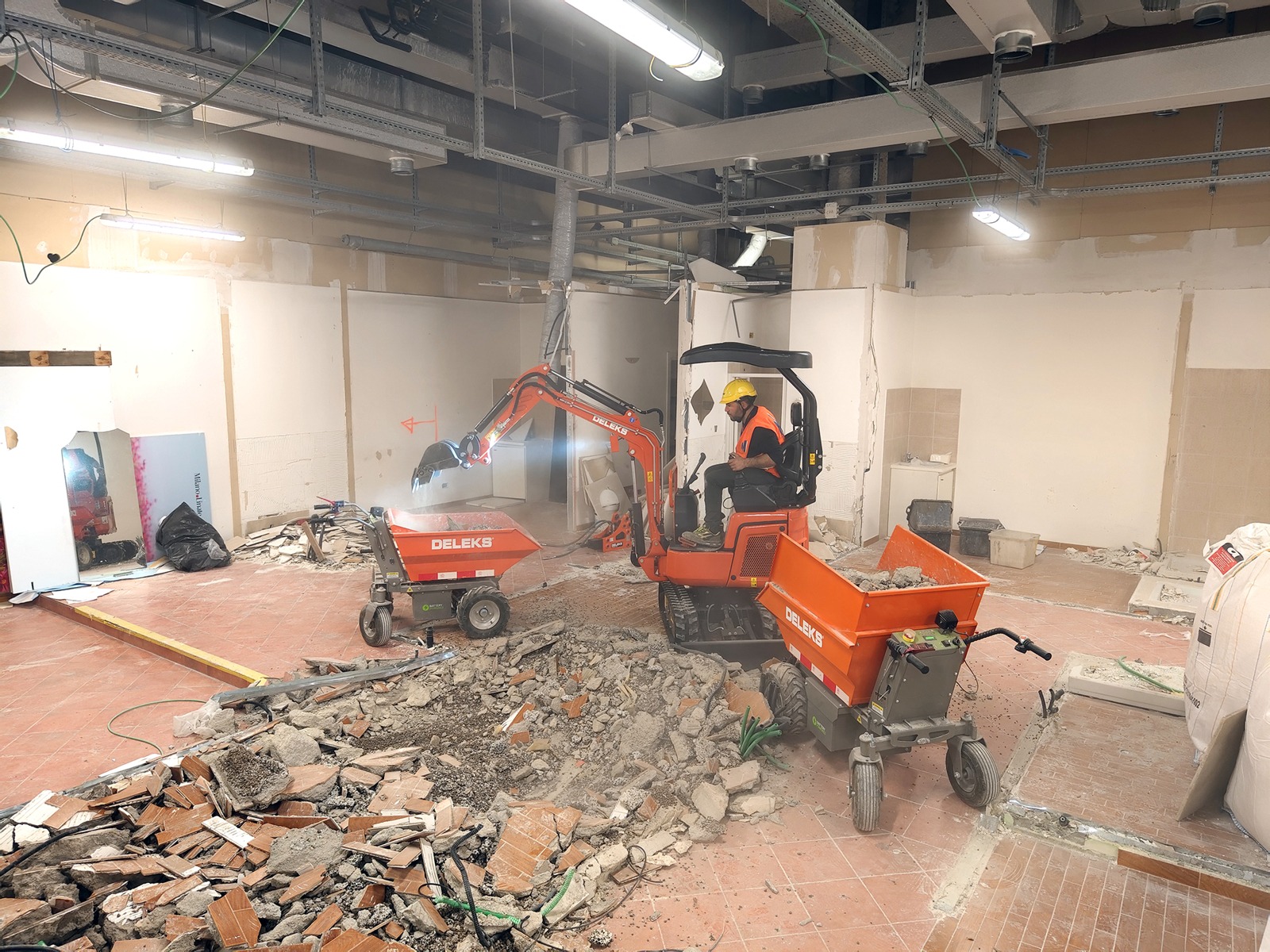 Töötaja juhib väikest punast lammutusekskavaatorit renoveeritavas hoones, mida ümbritsevad prahi, purunenud plaadid ning avatud seinad ja laejuhtmestik.