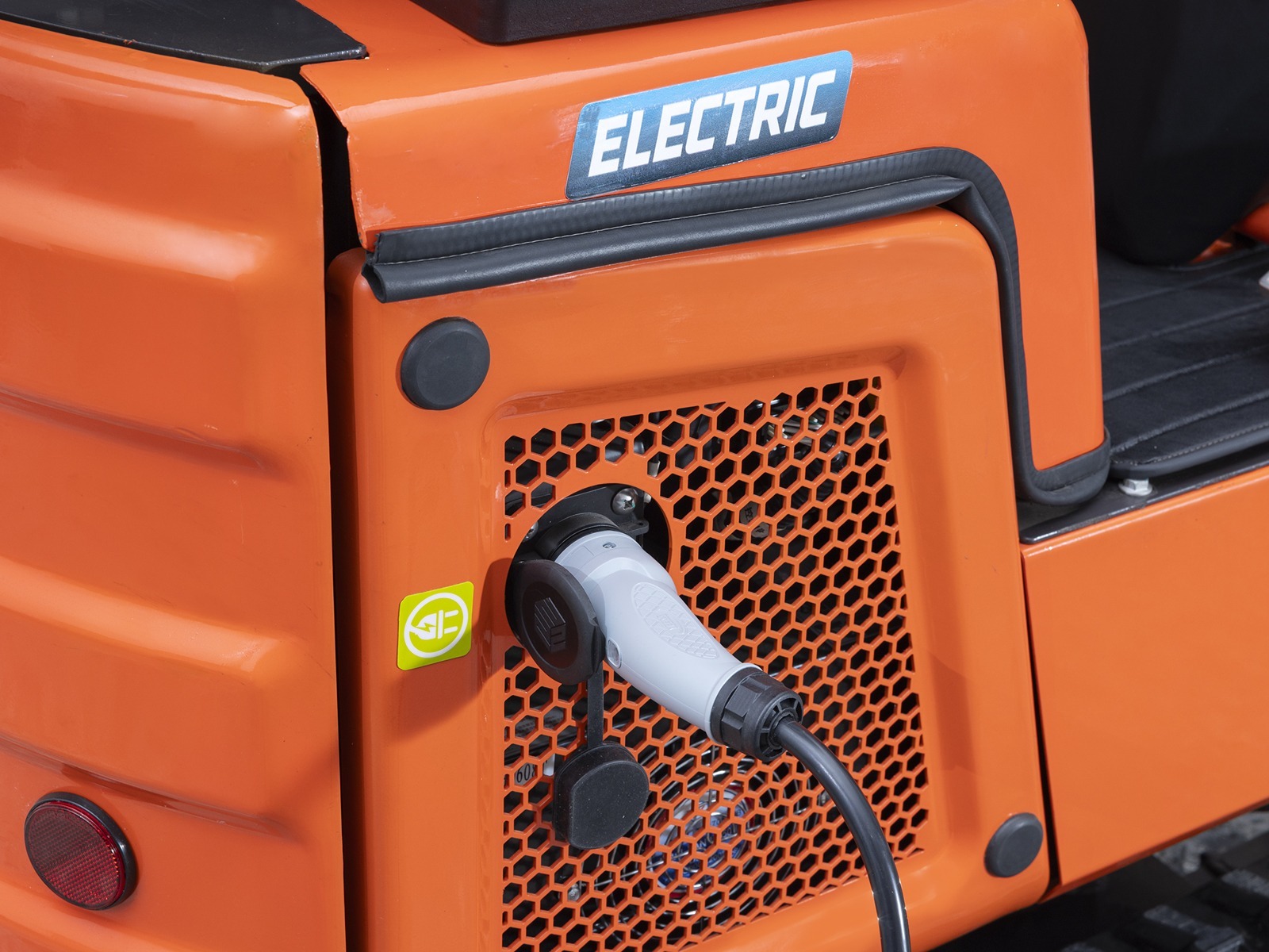 Lähivaade elektrilisest ehitusautost, millel on laadimiskaabel ühendatud laadimisporti, mille kohal on silt ELECTRIC ja taustal oranž perforeeritud paneel.