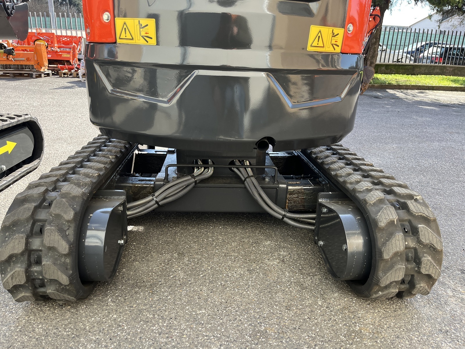 Lähivaade asfaldile pargitud kompaktekskavaatori tagumisest alumisest osast, millel on näha kummirööpad, metallraam, hüdraulikajuhtmed ja hoiatuskleebised.
