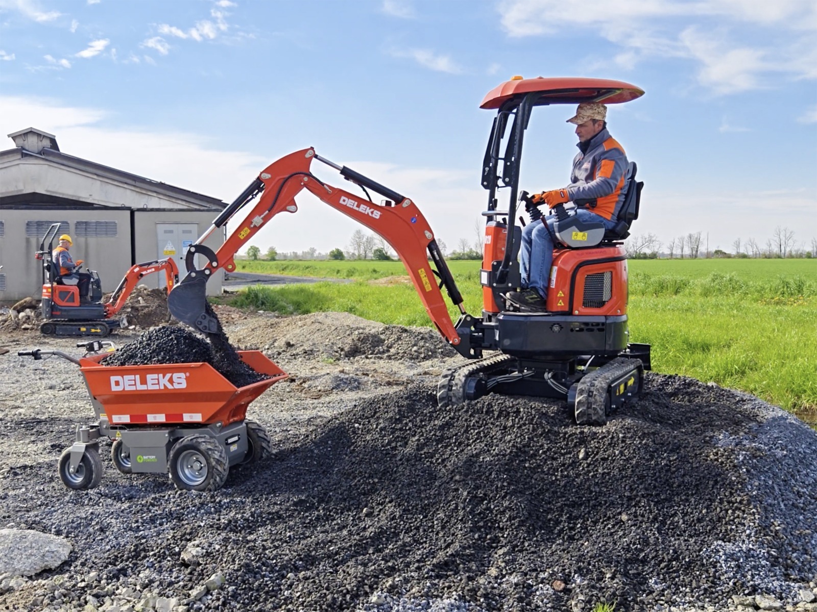 Inimene juhib väikest oranži värvi ekskavaatorit, mis liigutab kruusa lähedalasuvasse kompaktsesse kallurautosse. Taustal on hoone lähedal näha veel üks töötaja. Stseen on päikesepaistelisel päeval õues, mida ümbritsevad rohelised põllud.