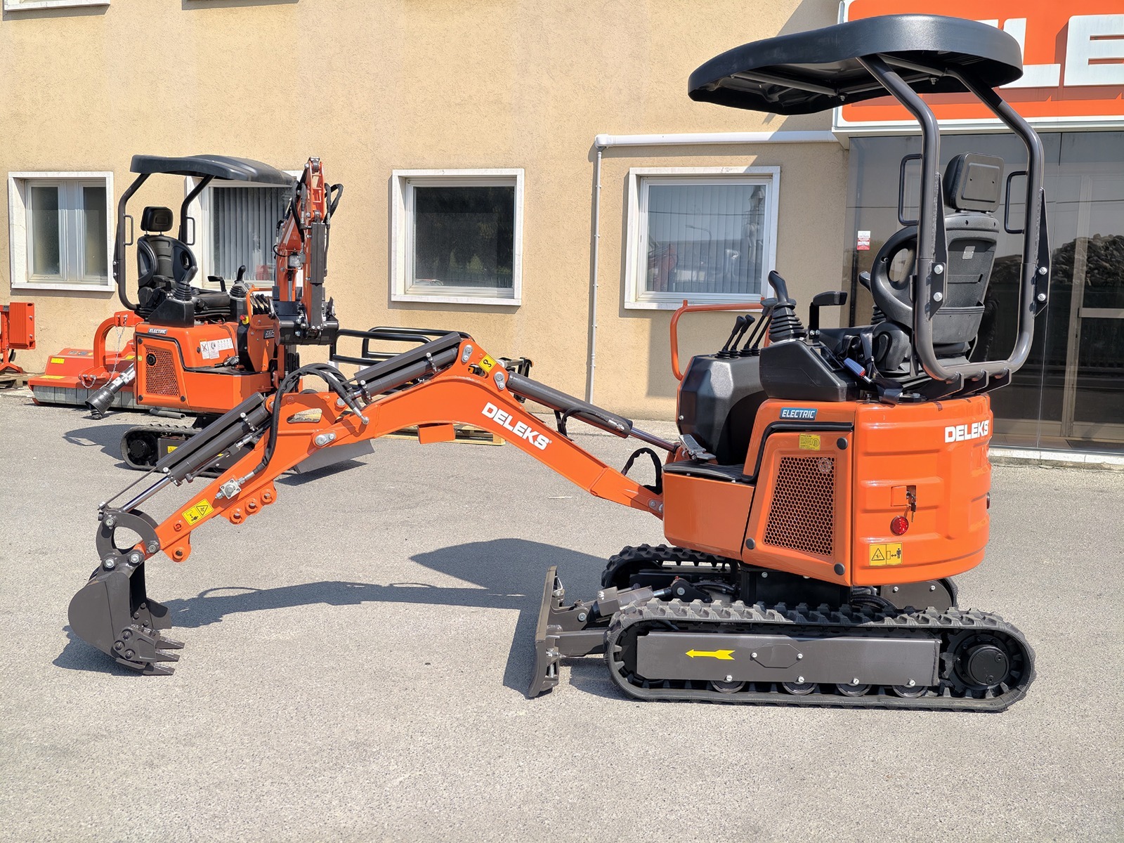 Oranžikas kompaktne ekskavaator, millel on kummirajad ja esitera, on pargitud sillutatud pinnale akendega beeži hoone ette. Taustal on näha veel üks sarnane masin.