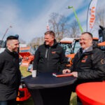 Kolm mustades jakkides meest seisavad ja vestlevad Maamessil 2026 välitingimustes kõrge ümmarguse laua ümber, taustal on päikesepaistelisel päeval näha oranžid traktorid ja Kone Agro bännerid.