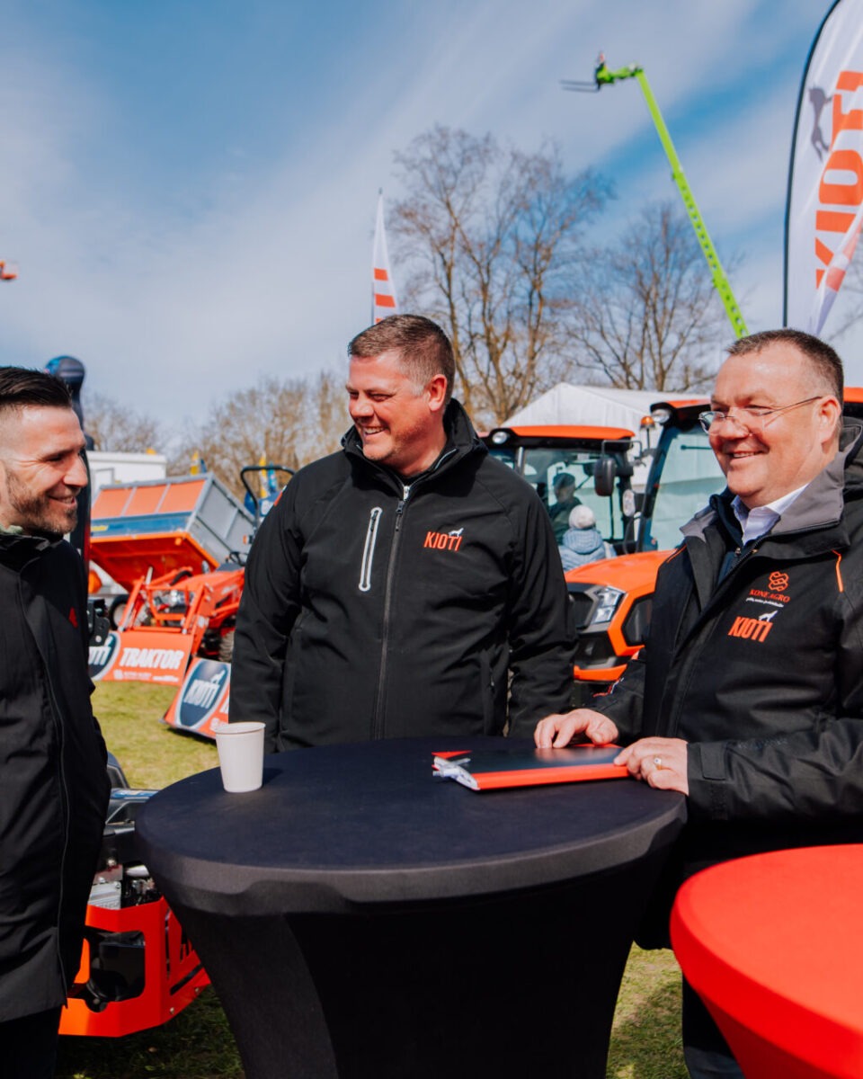 Kolm mustades jakkides meest seisavad ja vestlevad Maamessil 2026 välitingimustes kõrge ümmarguse laua ümber, taustal on päikesepaistelisel päeval näha oranžid traktorid ja Kone Agro bännerid.